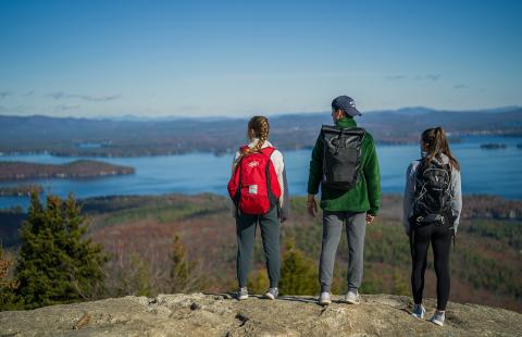 Three young people with backpacks stand on a mountain top over looking Lake Winnipesaukee