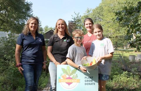 Three adults and two children in garden, holding produce and Plant a Row sign