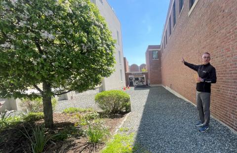 A man standing in a wide alley between brick buildings points to a small landscaped plot between buildings.